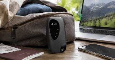 Routeur mobile TP-Link M7000 posé sur une table à côté d’un ordinateur portable, d’un smartphone et d’un sac de voyage